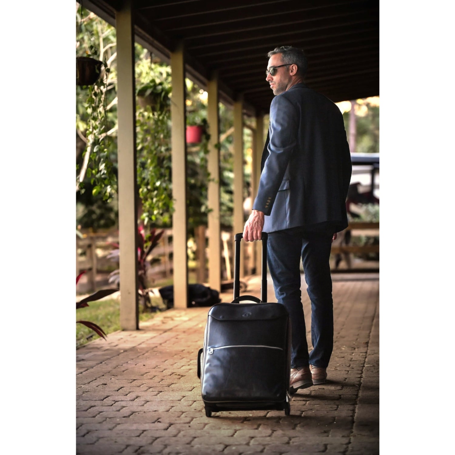 Man wearing a navy blazer, jeans, and sunglasses, pulling a black leather trolley suitcase along a wooden outdoor walkway surrounded by lush greenery