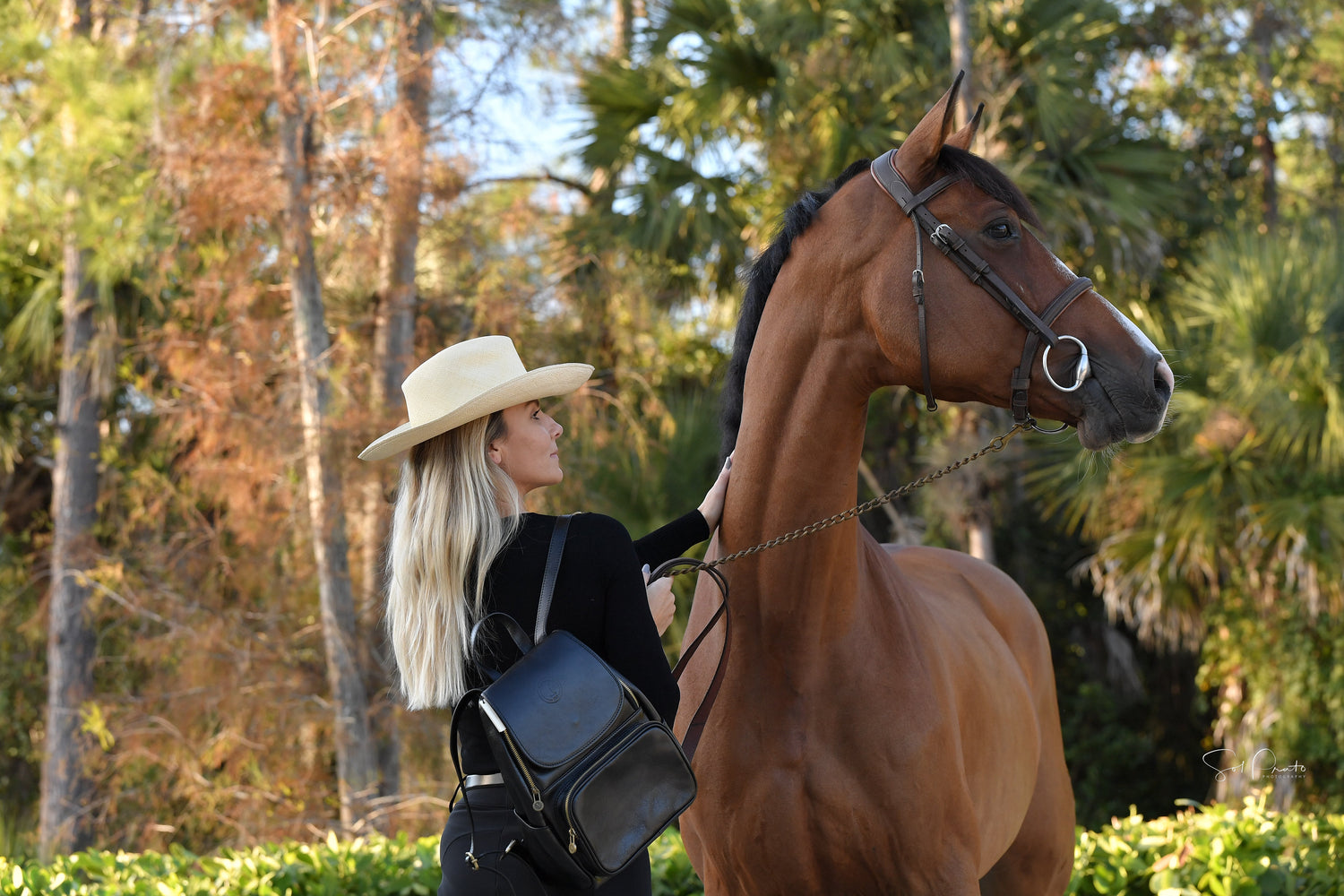 "Blonde woman wearing a straw hat and black leather backpack, gently holding a brown horse with a lush green forest in the background."